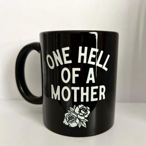 May include: Black ceramic coffee mug with white text that reads "One Hell of a Mother" and a white rose design.