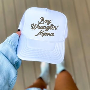 May include: White trucker hat with a gold rope-style embroidered design that says "Boy Wranglin' Mama".