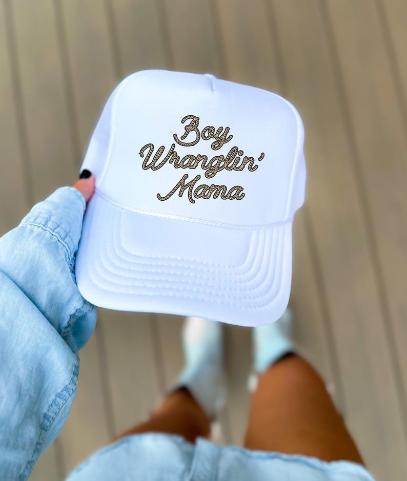 May include: White trucker hat with a gold rope-style embroidered design that says "Boy Wranglin' Mama".