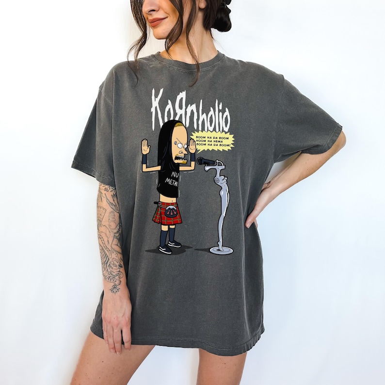 May include: A gray t-shirt with a cartoon image of a character from the animated series "Beavis and Butt-Head" wearing a kilt and holding a microphone. The text "Kornholio" and "Nu Metal" are printed on the shirt. The character is singing into the microphone and the text "BOOM NA DA BOOM BOOM NA DA BOOM BOOM NA DA BOOM" is printed in a speech bubble above the character.