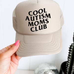 May include: A beige trucker hat with black text that reads "COOL AUTISM MOMS CLUB".