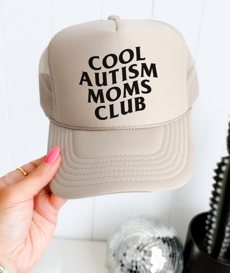 May include: A beige trucker hat with black text that reads "COOL AUTISM MOMS CLUB".