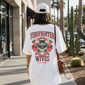 May include: White t-shirt with a graphic design on the back featuring the text "FIREFIGHTER WIVES CLUB" above a skull wearing a firefighter helmet. The design includes crossed axes and red accents. The person is wearing a white cap and carrying a brown purse.