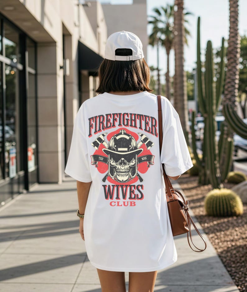 May include: White t-shirt with a graphic design on the back featuring the text "FIREFIGHTER WIVES CLUB" above a skull wearing a firefighter helmet. The design includes crossed axes and red accents. The person is wearing a white cap and carrying a brown purse.