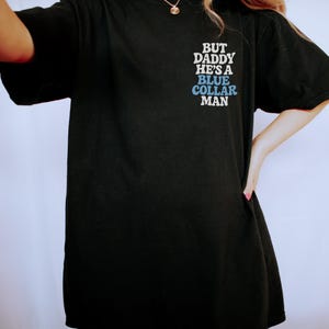 May include: Black t-shirt with the text "BUT DADDY HE'S A BLUE COLLAR MAN" in white and blue lettering. The shirt is a casual style, likely made of cotton or a similar fabric.