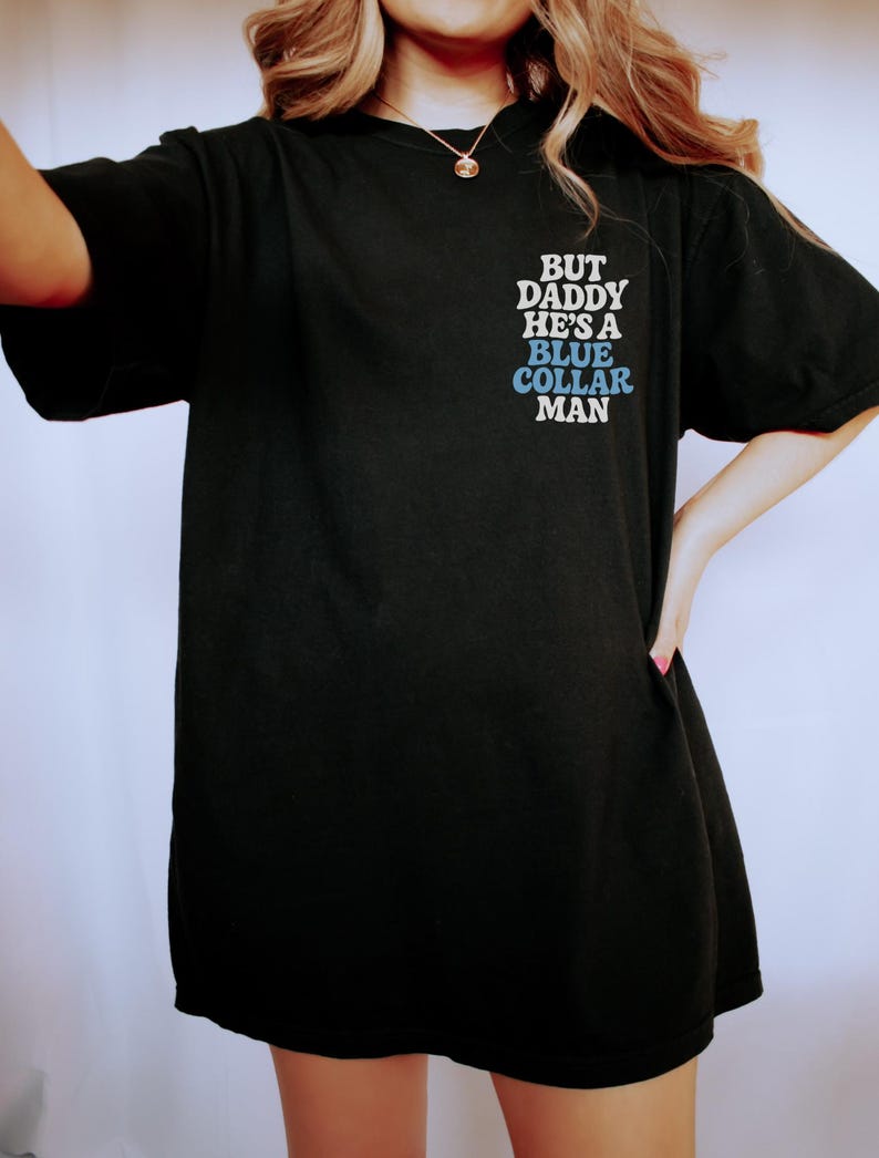 May include: Black t-shirt with the text "BUT DADDY HE'S A BLUE COLLAR MAN" in white and blue lettering. The shirt is a casual style, likely made of cotton or a similar fabric.