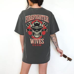 May include: Dark gray t-shirt with a graphic on the back featuring a firefighter's helmet, skull, and crossed axes, with the words "Firefighter Wives Club" in red and white.