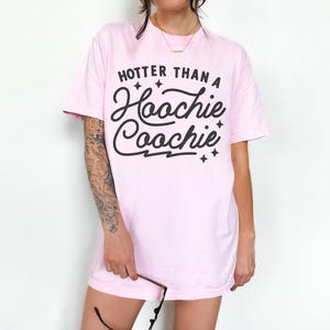 Puede incluir: Camiseta rosa claro con la frase "Hotter Than A Hoochie Coochie" en negro. La camiseta tiene mangas cortas y cuello redondo. La persona sostiene gafas.