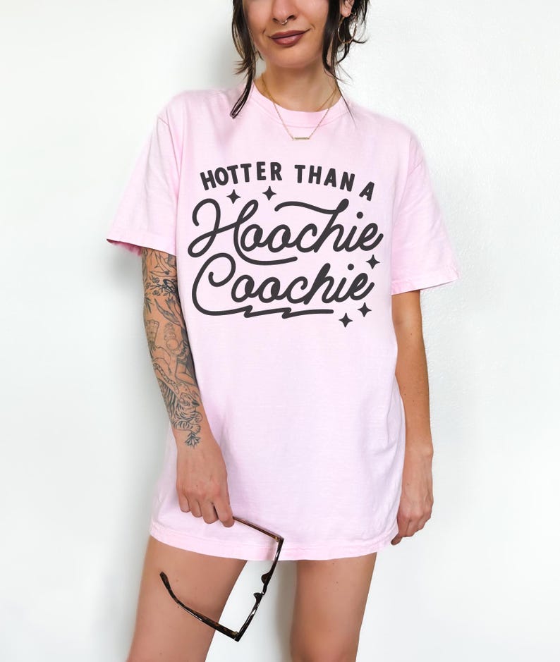 Puede incluir: Camiseta rosa claro con la frase "Hotter Than A Hoochie Coochie" en negro. La camiseta tiene mangas cortas y cuello redondo. La persona sostiene gafas.