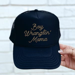 May include: Black trucker hat with a gold embroidered design that says "Boy Wranglin' Mama".