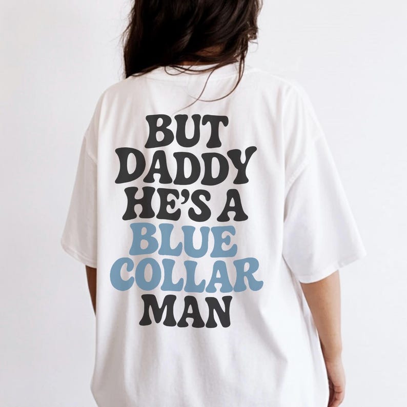 May include: White t-shirt with the text "BUT DADDY HE'S A BLUE COLLAR MAN" in black and blue lettering. The shirt has short sleeves and a relaxed fit. The text is centered on the back of the shirt.