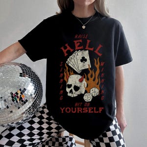 Può includere: Maglietta nera con una grafica di uno scheletro con corna, carte da gioco e fiamme. Il testo sulla maglietta recita "Raise Hell Sinning Winning Bet on Yourself".
