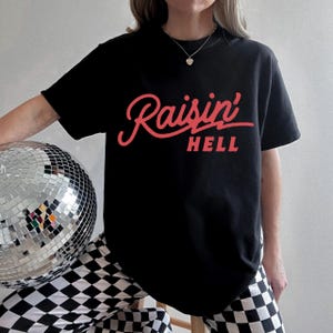 Puede incluir: Una camiseta negra con un gráfico rojo que dice "Raisin' Hell" en una fuente cursiva. La persona que lleva la camiseta también lleva pantalones a cuadros blanco y negro.