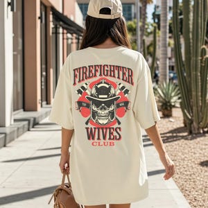 May include: Cream-colored oversized t-shirt with a graphic on the back. The graphic features a firefighter's helmet, crossed axes, and a skull, with the words "Firefighter Wives Club" in red and black. The person is wearing a beige baseball cap.