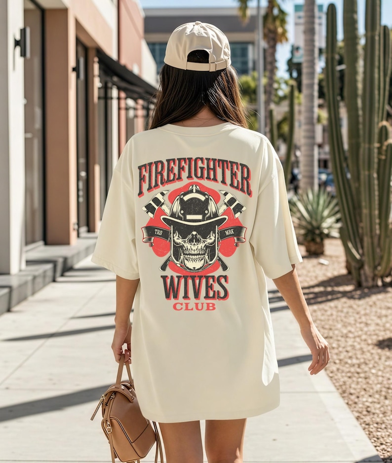 May include: Cream-colored oversized t-shirt with a graphic on the back. The graphic features a firefighter's helmet, crossed axes, and a skull, with the words "Firefighter Wives Club" in red and black. The person is wearing a beige baseball cap.