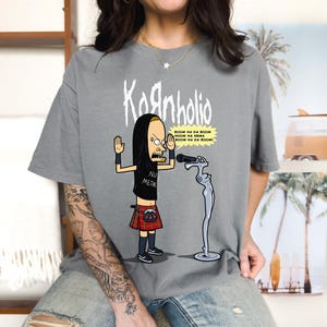 May include: Gray t-shirt with a cartoon image of a man wearing a kilt and holding a microphone. The text "Kornholio" and "BOOM NA DA BOOM NOOM HA NEMA BOOM NA DA BOOM!" is printed on the shirt. The man is wearing a black hooded shirt and the text "NU METAL" is printed on his shirt.