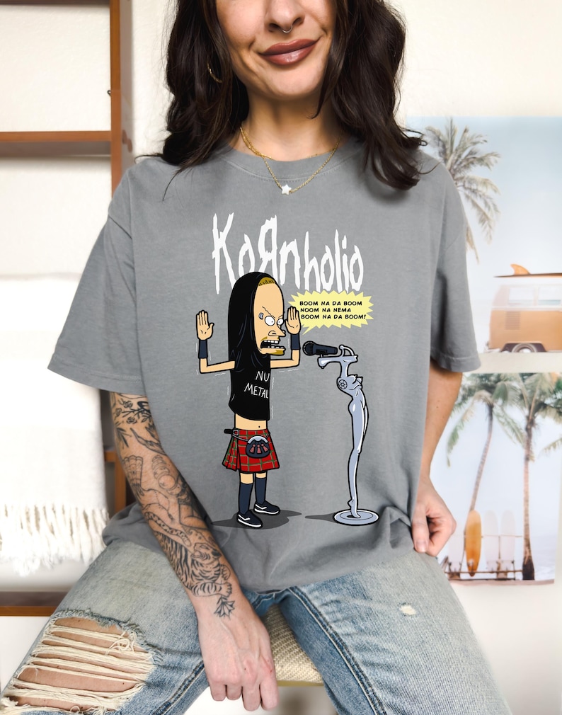 May include: Gray t-shirt with a cartoon image of a man wearing a kilt and holding a microphone. The text "Kornholio" and "BOOM NA DA BOOM NOOM HA NEMA BOOM NA DA BOOM!" is printed on the shirt. The man is wearing a black hooded shirt and the text "NU METAL" is printed on his shirt.