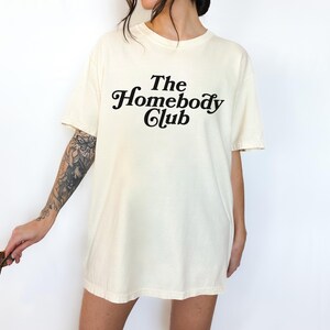 Może przedstawiać: Kremowa koszulka z napisem "The Homebody Club" w czarnym, ozdobnym pismie.