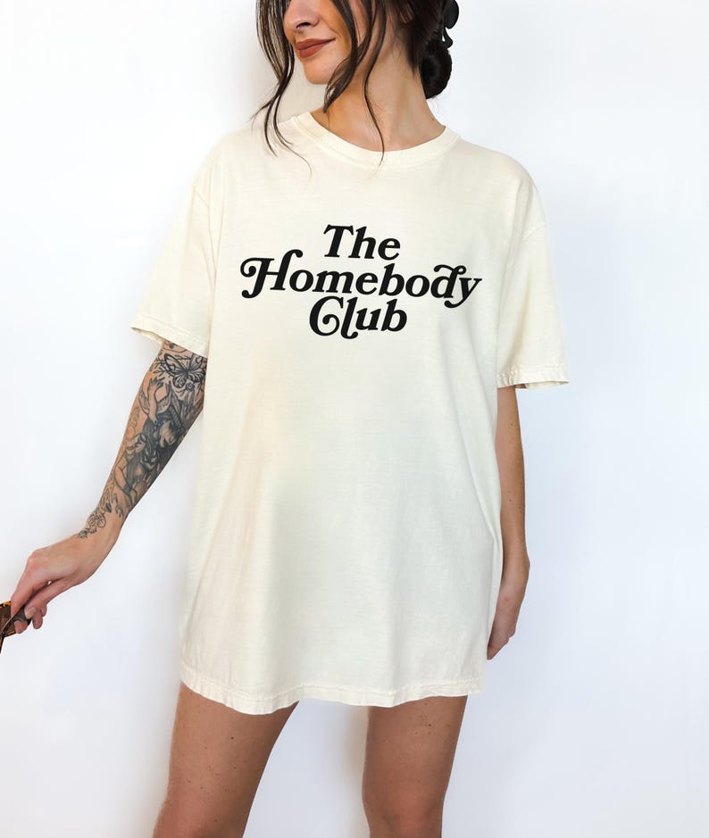 Może przedstawiać: Kremowa koszulka z napisem "The Homebody Club" w czarnym, ozdobnym pismie.