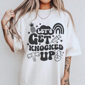 Op de afbeelding: Crèmekleurig T-shirt met de zwarte tekst "LET'S GET KNOCKED UP", samen met illustraties van een ananas, sterren, een regenboog, een spuit, een baarmoeder en een handgebaar.