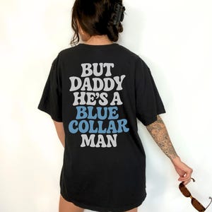May include: Black t-shirt with the text "BUT DADDY HE'S A BLUE COLLAR MAN" in white and blue lettering. The shirt is a casual style, with short sleeves.