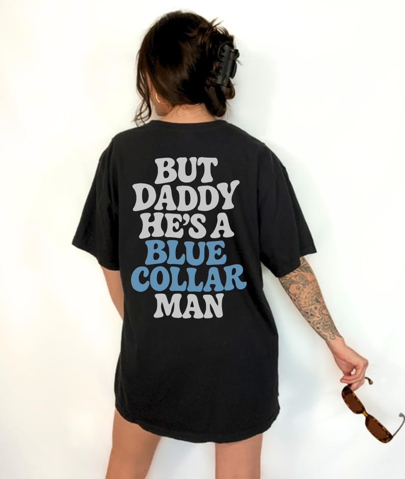 May include: Black t-shirt with the text "BUT DADDY HE'S A BLUE COLLAR MAN" in white and blue lettering. The shirt is a casual style, with short sleeves.
