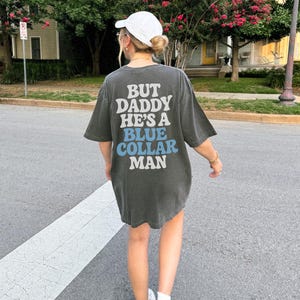 May include: Dark gray oversized t-shirt with the text "BUT DADDY HE'S A BLUE COLLAR MAN" in white and blue lettering. The shirt is worn with a white cap and sneakers. The image is taken outdoors on a sunny day.