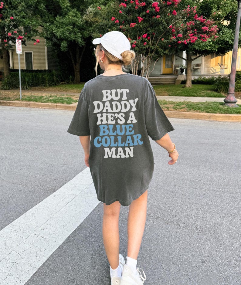 May include: Dark gray oversized t-shirt with the text "BUT DADDY HE'S A BLUE COLLAR MAN" in white and blue lettering. The shirt is worn with a white cap and sneakers. The image is taken outdoors on a sunny day.