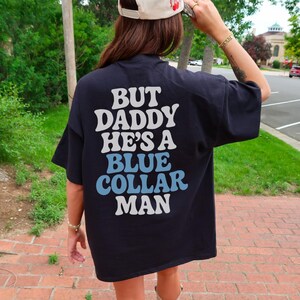 May include: Black t-shirt with the text "BUT DADDY HE'S A BLUE COLLAR MAN" in white and light blue lettering. The shirt is worn with a cream-colored baseball cap with a red cherry design.