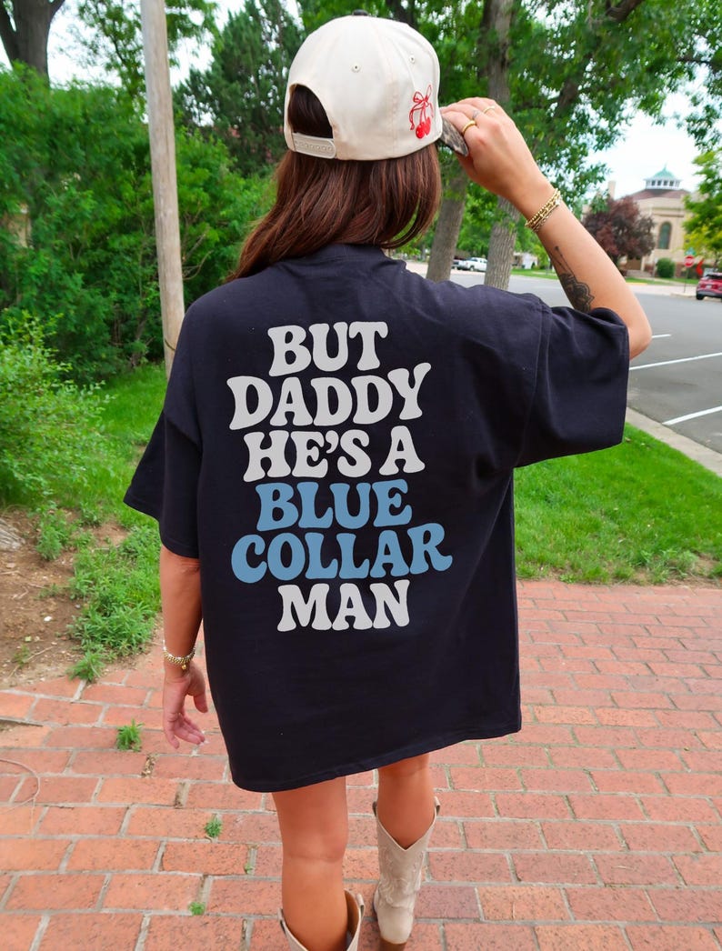 May include: Black t-shirt with the text "BUT DADDY HE'S A BLUE COLLAR MAN" in white and light blue lettering. The shirt is worn with a cream-colored baseball cap with a red cherry design.