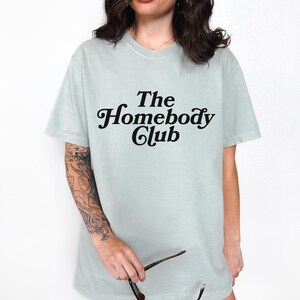 Może przedstawiać: Jasnozielony t-shirt z napisem "The Homebody Club" w czarnym retro foncie.