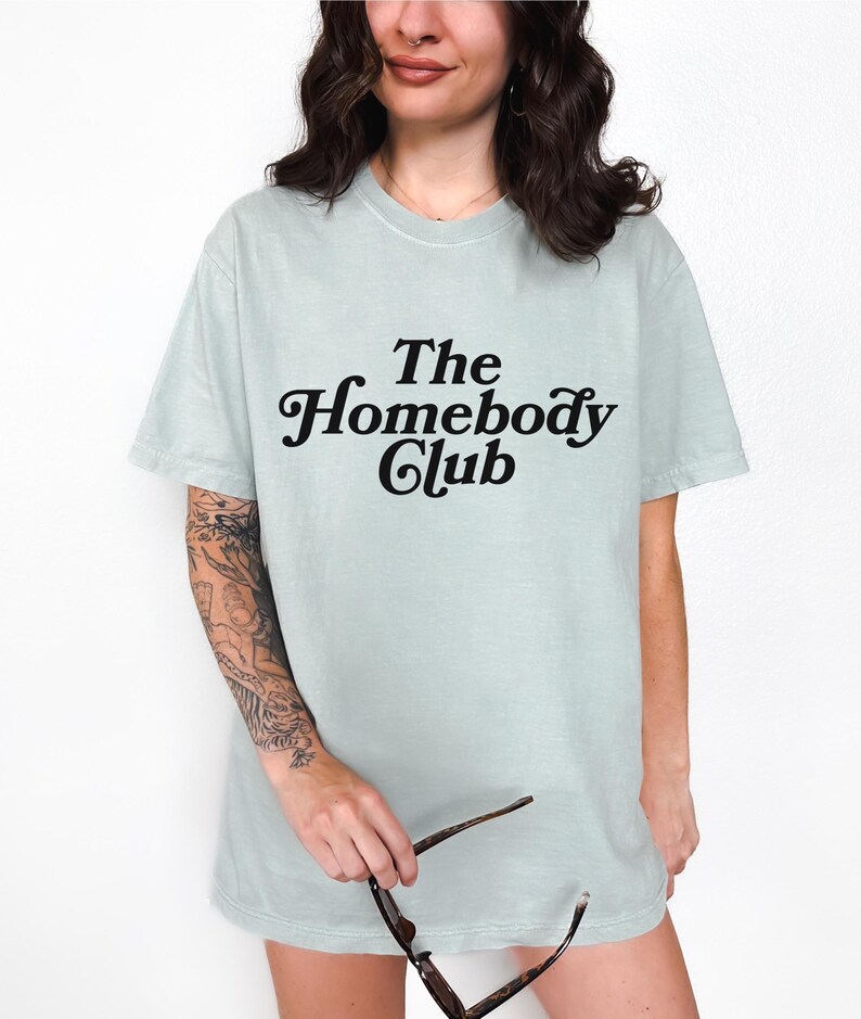 Może przedstawiać: Jasnozielony t-shirt z napisem "The Homebody Club" w czarnym retro foncie.