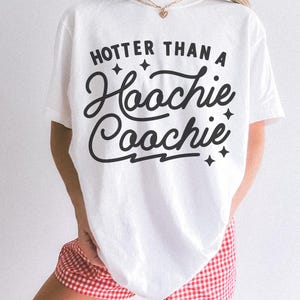 Puede incluir: Camiseta blanca con la frase "Hotter Than A Hoochie Coochie" en letras negras. La camiseta se combina con pantalones cortos a cuadros rojos y blancos. El estilo general es informal y relajado.