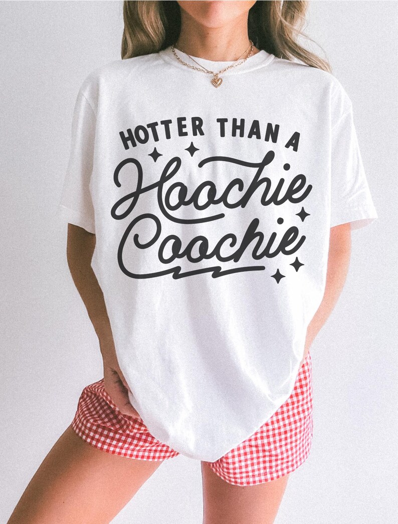 Puede incluir: Camiseta blanca con la frase "Hotter Than A Hoochie Coochie" en letras negras. La camiseta se combina con pantalones cortos a cuadros rojos y blancos. El estilo general es informal y relajado.