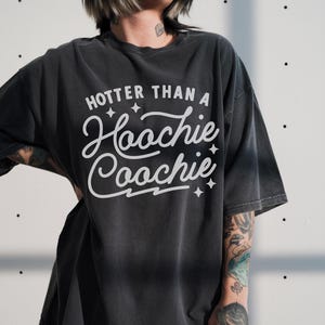 Puede incluir: Camiseta gris oscuro con la frase "Hooter Than a Hoochie Coochie" en blanco. La camiseta tiene un ajuste holgado y mangas cortas. El texto est&aacute; centrado en la parte delantera, con detalles de estrellas.