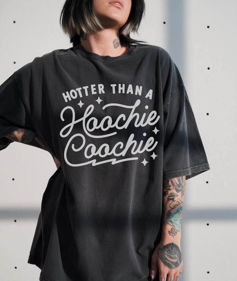 Puede incluir: Camiseta gris oscuro con la frase "Hooter Than a Hoochie Coochie" en blanco. La camiseta tiene un ajuste holgado y mangas cortas. El texto est&aacute; centrado en la parte delantera, con detalles de estrellas.