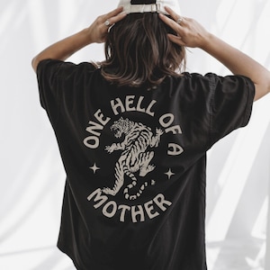 May include: Black t-shirt with a white tiger graphic and the text "ONE HELL OF A MOTHER". The design includes stars. The shirt is a casual style.
