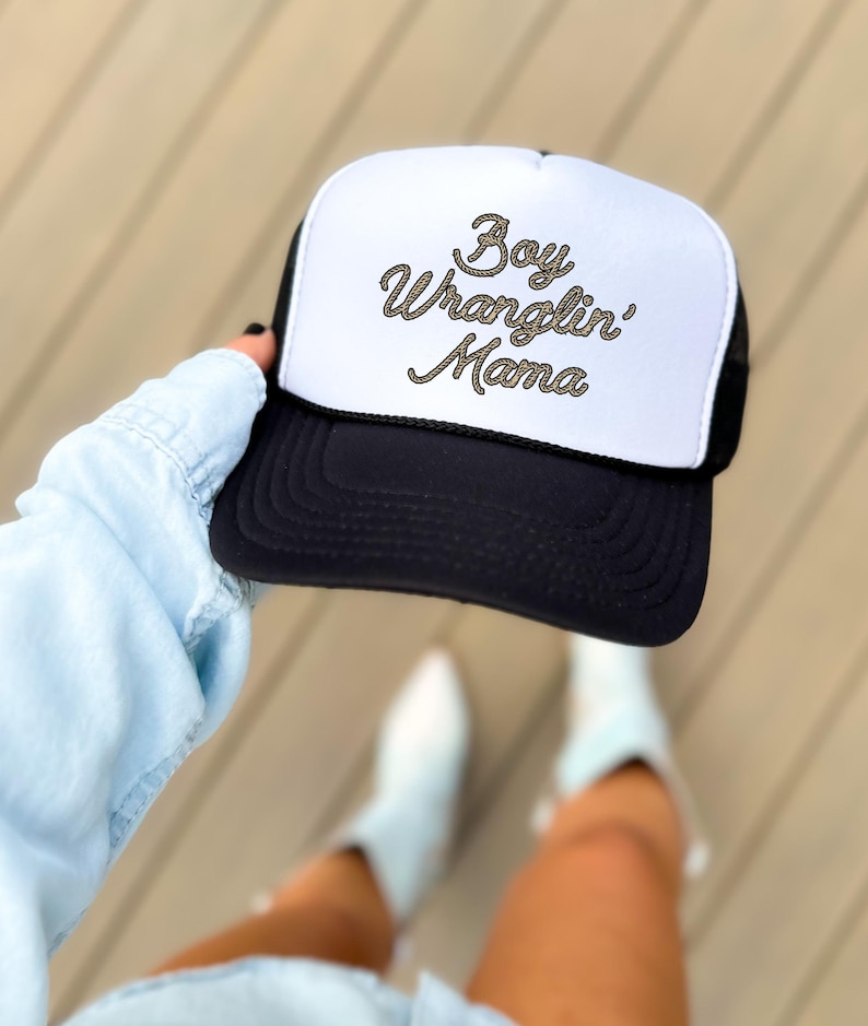 May include: A black and white trucker hat with a white mesh back. The hat has gold rope lettering that reads "Boy Wranglin' Mama".