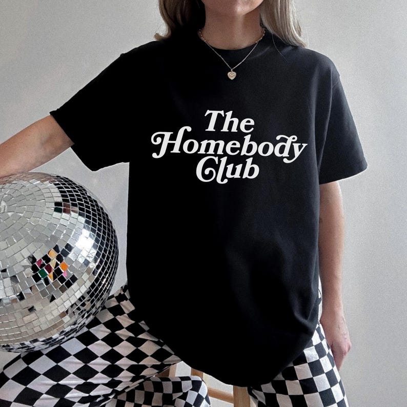 Może przedstawiać: Czarny T-shirt z białym napisem "The Homebody Club" w retro czcionce. Osoba nosząca T-shirt trzyma srebrną kulę dyskotekową.