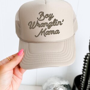 May include: A beige trucker hat with a brown rope-style embroidered design that says "Boy Wranglin' Mama".
