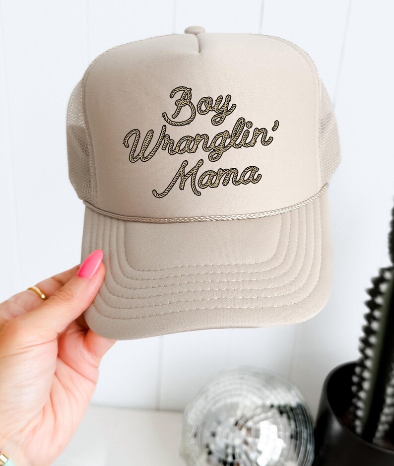 May include: A beige trucker hat with a brown rope-style embroidered design that says "Boy Wranglin' Mama".