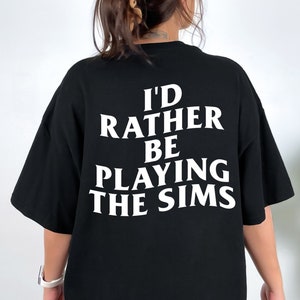 Puede incluir: Camiseta negra con texto blanco que dice "I'd rather be playing the Sims".