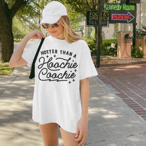 Puede incluir: Camiseta blanca con el texto "HOTTER THAN A Hoochie Coochie" en fuente negra. La persona lleva una gorra blanca y gafas de sol.