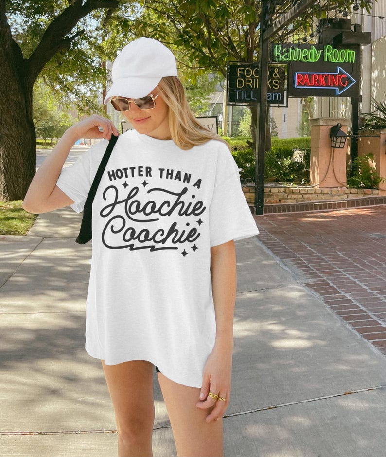 Puede incluir: Camiseta blanca con el texto "HOTTER THAN A Hoochie Coochie" en fuente negra. La persona lleva una gorra blanca y gafas de sol.