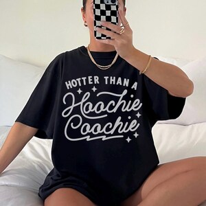 Puede incluir: Camiseta negra con la frase blanca "Hooter Than a Hoochie Coochie". La camiseta tiene mangas cortas y un corte holgado. La imagen muestra la camiseta puesta.