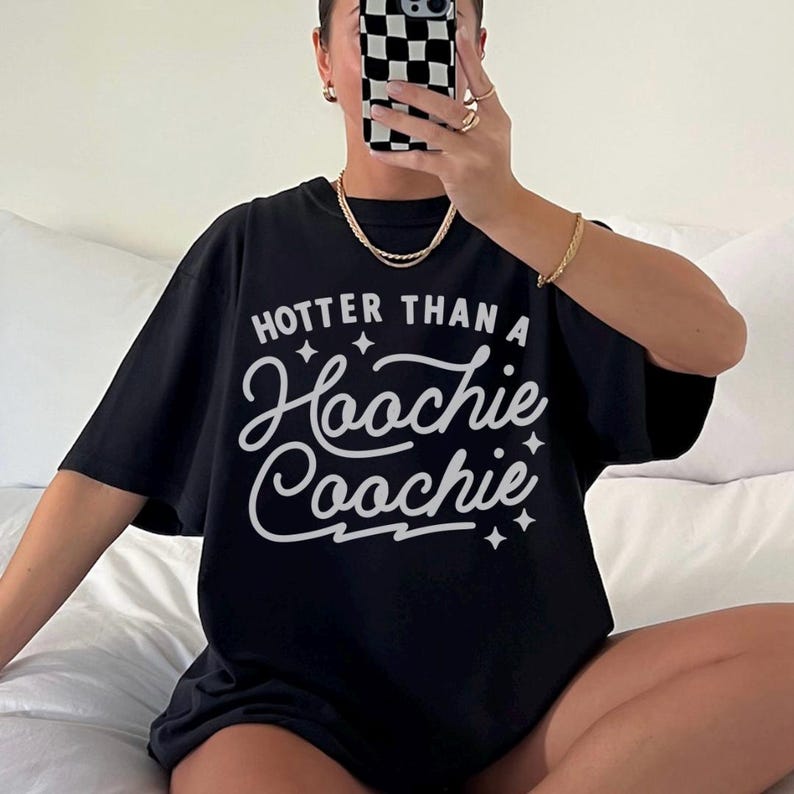 Puede incluir: Camiseta negra con la frase blanca "Hooter Than a Hoochie Coochie". La camiseta tiene mangas cortas y un corte holgado. La imagen muestra la camiseta puesta.