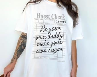 以下が含まれることがあります： 黒い「Be your own daddy make your own sugar」というゲストチェックのグラフィックがプリントされた白いTシャツです。