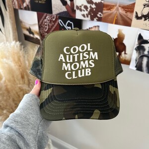 May include: A green camouflage trucker hat with a white mesh back. The front of the hat has the text "COOL AUTISM MOMS CLUB" in white.