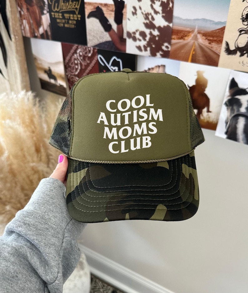 May include: A green camouflage trucker hat with a white mesh back. The front of the hat has the text "COOL AUTISM MOMS CLUB" in white.