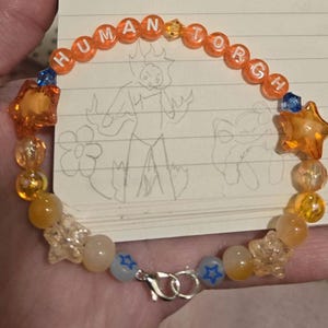 Op de afbeelding: Een kralenarmband met het woord "HUMAN TORCH" gespeld in oranje letterkralen. De armband heeft oranje stervormige kralen, heldere stervormige kralen en andere oranje en gele kralen. Een zilveren sluiting zet de armband vast.